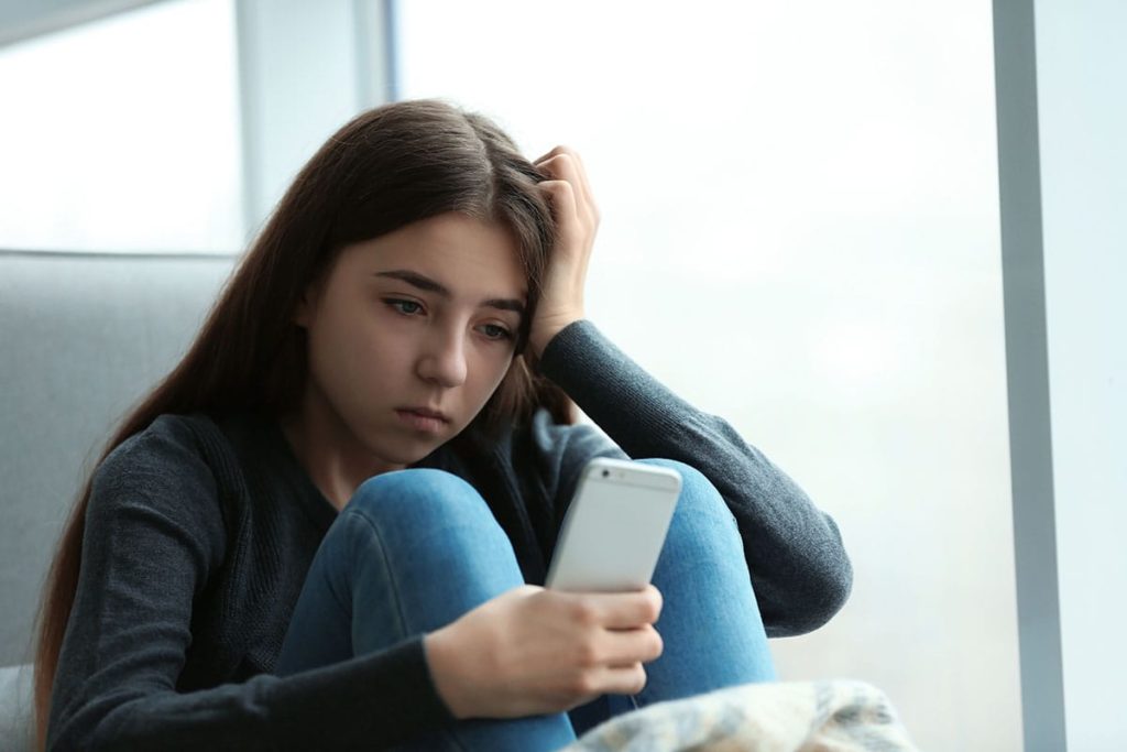 teenage girl staring bleakly at her smartphone wondering what is cyberbullying and how to stop it