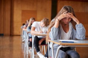 image of students at desks in a classroom with female student holding her head in her hands and displaying common anxiety triggers