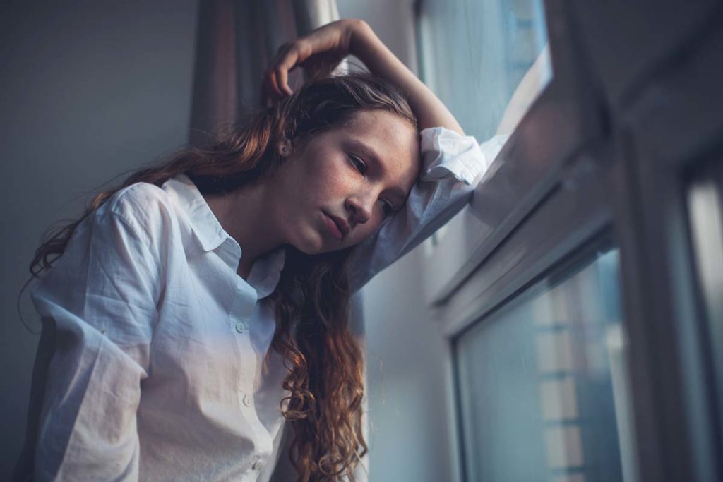 teenage girl in darkened bedroom leaning against window frame and coping with depression