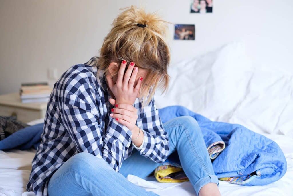 a teen with antisocial behavior sits on a bed with their face in their hands