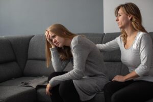 mother seated on couch reaching out a hand to comfort her daughter wanting to know how o help teens struggling with grief